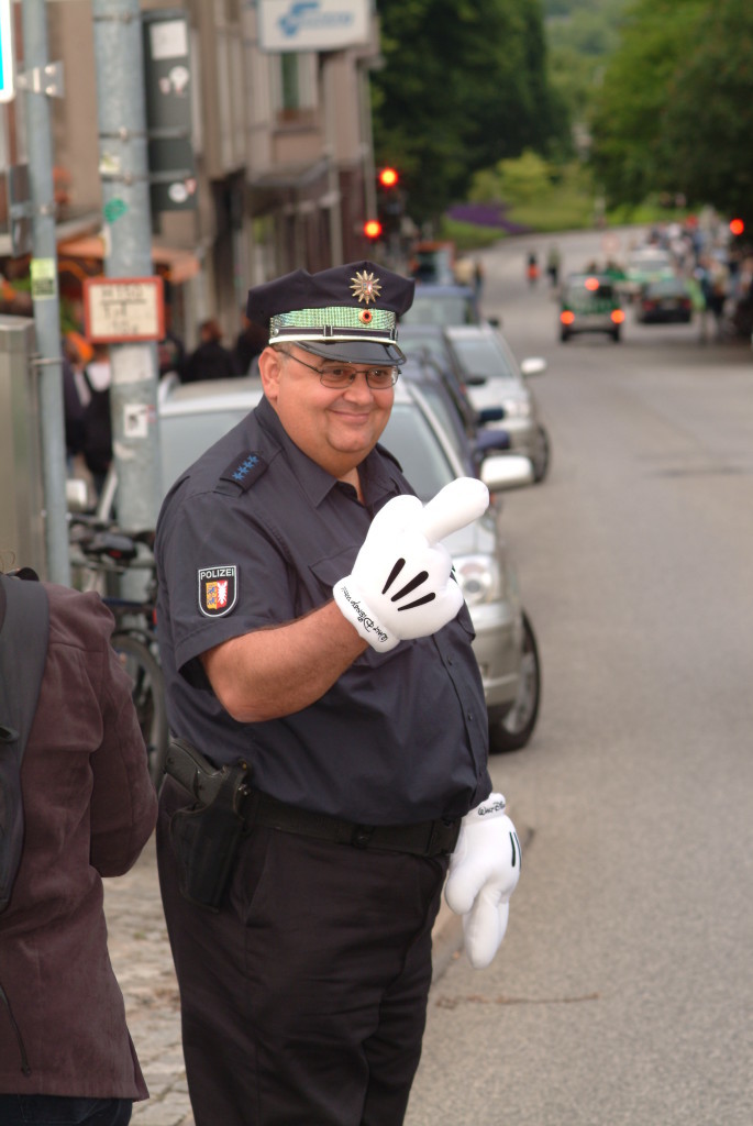 Hier kommen wir in einen absoluten Grenzbereich. Dieser Polizist hat den Verkehr geregelt anlässlich des Besuchs hochrangiger Politiker auf der Kieler Woche.Eigentlich darffst Du das Foto ohne seine Genehmigung nicht veröffentlichen. Nun spielen hier besondere Aspekte eine Rolle. Der Polizist hat sich mit den Handschuhen "verkleidet", durch die Uniform erweckt er in Kombination mit den Handschuhen Aufmerksamkeit. Es gibt neben diesem Foto weitere Aufnahmen, wo er sichtbar mit anderen Personen für Fotos posiert. Man nennt dies auch "konkludentes" Verhalten. Dennihm muss bewusst sein, dass er fotografiert wird und diese Fotos ggf.auch mal veröffentlicht werden. Sollte er gegen eine Veröffentlichung vorgehen, würde es ihm schwerfallen, dies zu begründen.In Sume also ein kalkulierbares Risiko so ein Foto zu veröffentllichen.