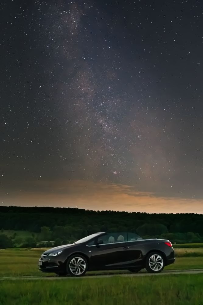 Ein Cabriolet fährt auf einer malerischen Straße durch einen dunklen Wald, während die beeindruckende Milchstraße sich über dem klaren Nachthimmel erstreckt.